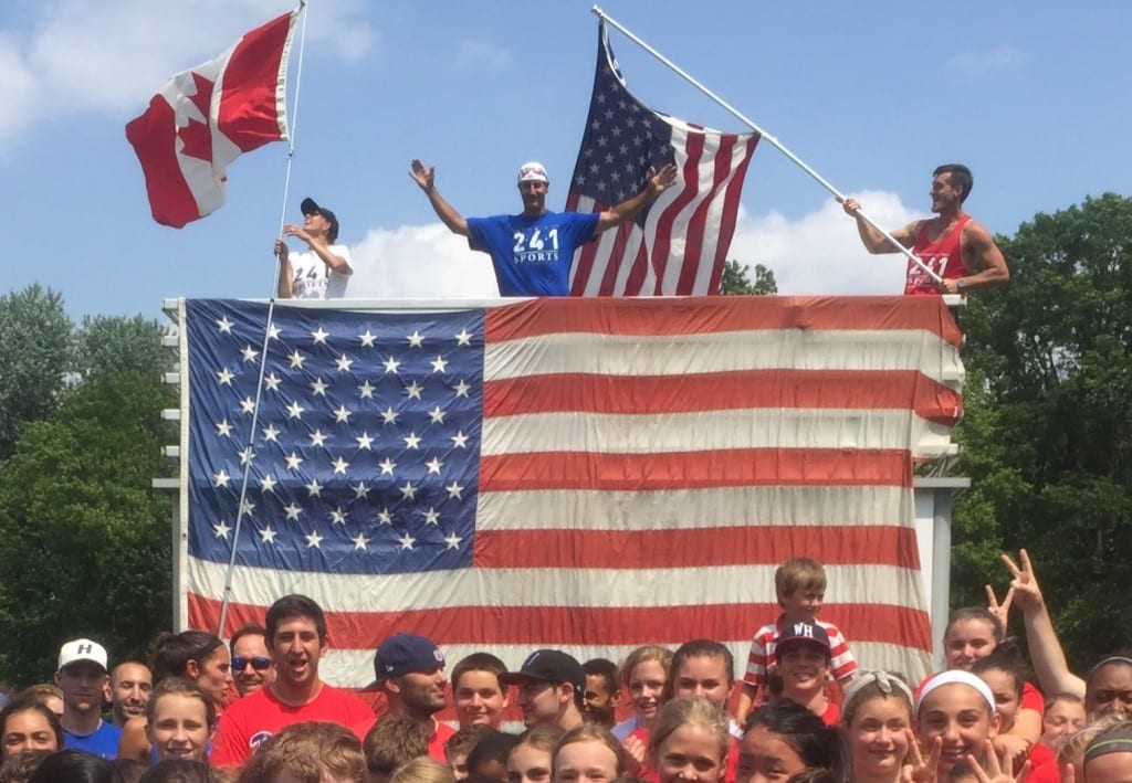 Ryan Radmanovich, arms raised,  gazes out over the American Flag, with all the campers in the foreground at the party celebrating his U.S. citizenship. Photo credit: Olivia Van Rye