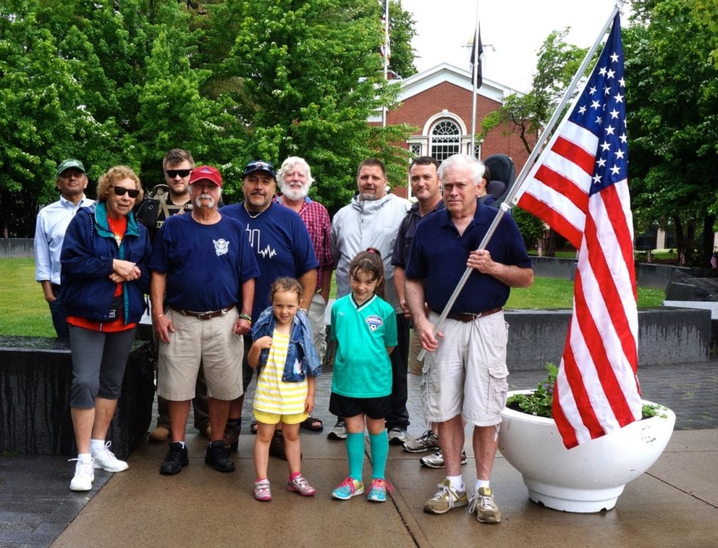 A group of veterans and supporters pay tribute to the fallen at the Connecticut Veterans Memorial in West Hartford after staging an informal parade along the Farmington Avenue sidewalk. Gerry Aldrich, holding the flag, organized the group that included his wife Susan (at left), Giovanni Seccareccia (center with dark blue cap), Joe Goldkamp (back row in red shirt), and Sean Caffyn (back row at far right with baby in backpack). Photo credit: Ronni Newton