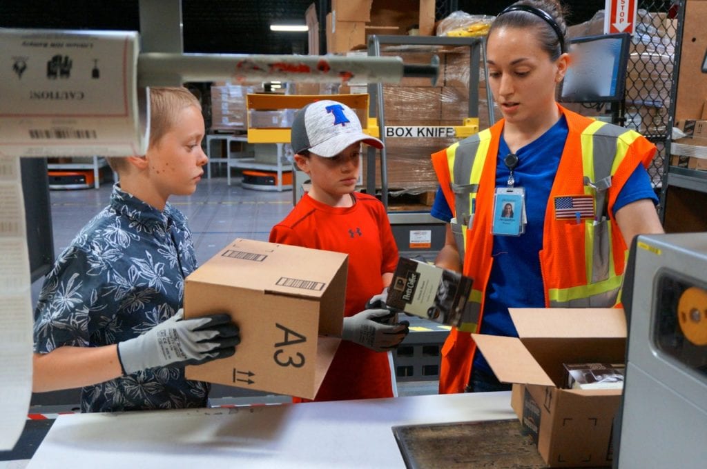 Britt Housum (left) and Alexander Hyams are instructed by Jackie of Amazon on how to place the product in the box. Photo credit: Ronni Newton